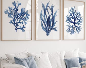 Op de afbeelding: Drie ingelijste wandkunstprints met blauwe aquarelillustraties van zeekoraal en zeewier op een witte achtergrond. De prints zijn ingelijst in licht houten lijsten en hangen in een horizontale rij boven een witte bank met blauwe kussens.