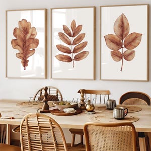 May include: Three framed wall art prints featuring watercolor illustrations of brown autumn leaves on a light background. The prints are displayed in light wood frames above a dining table with rattan chairs and various table decor items.
