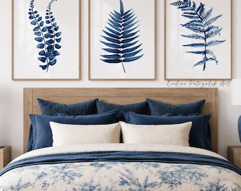 May include: A set of three framed wall art prints featuring dark blue botanical fern illustrations on white backgrounds, displayed above a bed with a blue and white floral patterned duvet, navy pillows, and two blue ceramic table lamps.