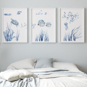 Può includere: Tre stampe incorniciate ad acquerello raffiguranti la vita marina. Le stampe raffigurano una tartaruga marina, pesci e una medusa, insieme ad alghe blu e altre piante acquatiche. L'opera d'arte è esposta sopra un letto con biancheria da letto bianca e una parete azzurra.