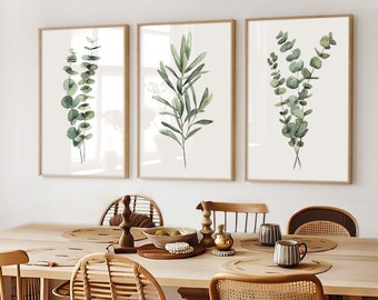 May include: Three framed botanical prints with watercolour eucalyptus and olive branch illustrations. The prints have light brown wooden frames and are displayed above a wooden dining table with woven chairs and decorative items.