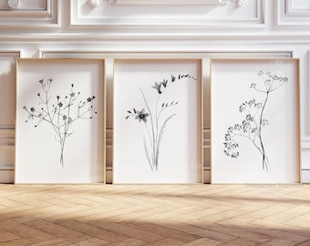 May include: Three framed wall art prints featuring black and white botanical illustrations of wildflowers. Each print displays a different delicate floral arrangement on a white background, housed in light-colored wooden frames.
