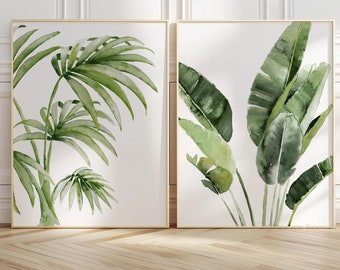 May include: Two framed wall art prints featuring watercolor illustrations of tropical plants. The left print displays a palm frond with slender green leaves, while the right print shows broad, elongated banana leaves in various shades of green.