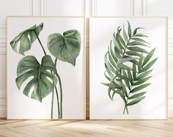 May include: Two framed watercolor prints of tropical leaves. The print on the left features a monstera leaf, while the print on the right features a palm leaf.