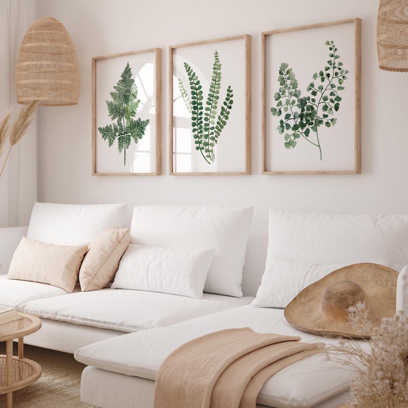 May include: Three framed watercolor prints of green ferns on a white wall above a white sectional sofa with a brown throw blanket and a brown hat on the sofa.