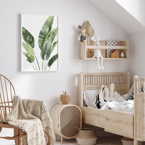 May include: A white wooden bed frame with a white and black patterned pillow and a white blanket. A wicker basket sits on the floor next to the bed. A white wicker nightstand with a woven rattan door sits next to the bed. A green watercolor painting of banana leaves hangs on the wall above a white rocking chair with a crocheted throw.