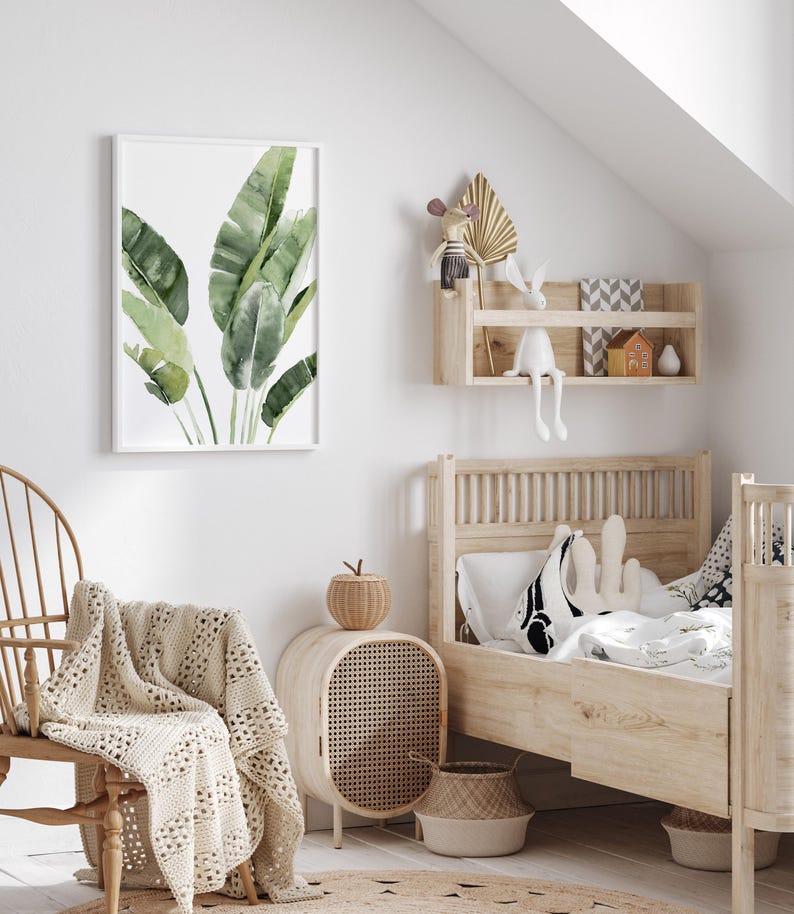 May include: A white wooden bed frame with a white and black patterned pillow and a white blanket. A wicker basket sits on the floor next to the bed. A white wicker nightstand with a woven rattan door sits next to the bed. A green watercolor painting of banana leaves hangs on the wall above a white rocking chair with a crocheted throw.