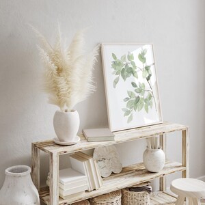 May include: A white wooden shelf unit with two shelves, featuring a white ceramic vase with dried pampas grass, a framed watercolor print of green leaves, a stack of books, and a ceramic vase with a white glaze.