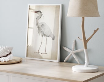 May include: A white dresser with a light wood top. A framed watercolor painting of a heron is on the top of the dresser. A white lamp with a beige lampshade and a wooden base is on the right side of the dresser. A white starfish is on the dresser in front of the lamp.