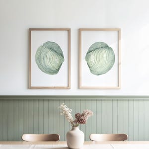 Op de afbeelding: Twee ingelijste aquarelprints van schelpen in groen- en wittinten. De prints zitten in licht houten frames en hangen aan een witte muur. Een vaas met droogbloemen staat op een tafel eronder.