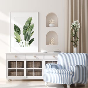 May include: A white cabinet with glass doors and wicker baskets. The cabinet has two drawers with wooden knobs. A blue and white striped armchair sits in front of the cabinet. A framed print of green leaves hangs on the wall above the cabinet.