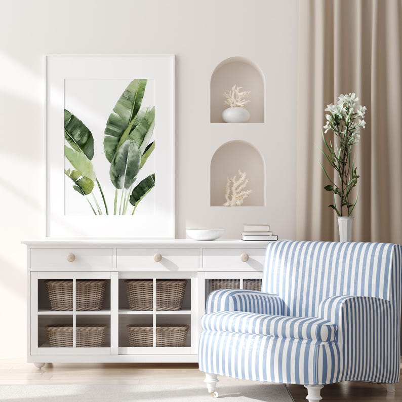 May include: A white cabinet with glass doors and wicker baskets. The cabinet has two drawers with wooden knobs. A blue and white striped armchair sits in front of the cabinet. A framed print of green leaves hangs on the wall above the cabinet.