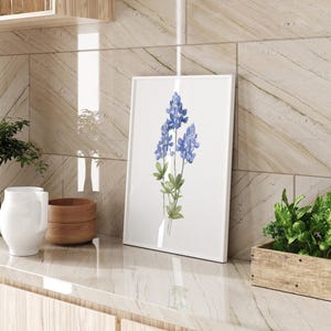 K&ouml;nnte beinhalten: Ein gerahmtes Aquarellbild von blauen Lupinen auf wei&szlig;em Hintergrund. Die Blumen stehen in einer Vase und sind von gr&uuml;nen Bl&auml;ttern umgeben.
