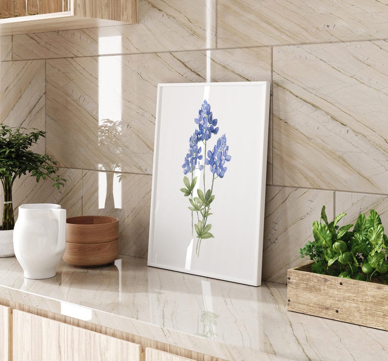 K&ouml;nnte beinhalten: Ein gerahmtes Aquarellbild von blauen Lupinen auf wei&szlig;em Hintergrund. Die Blumen stehen in einer Vase und sind von gr&uuml;nen Bl&auml;ttern umgeben.