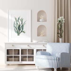 Könnte beinhalten: Ein weißer Schrank mit Glastüren und geflochtenen Körben. Ein blaugestreifter Sessel mit weißen Streifen steht vor dem Schrank. Ein gerahmtes Aquarellbild von grünem Seetang hängt an der Wand über dem Schrank.