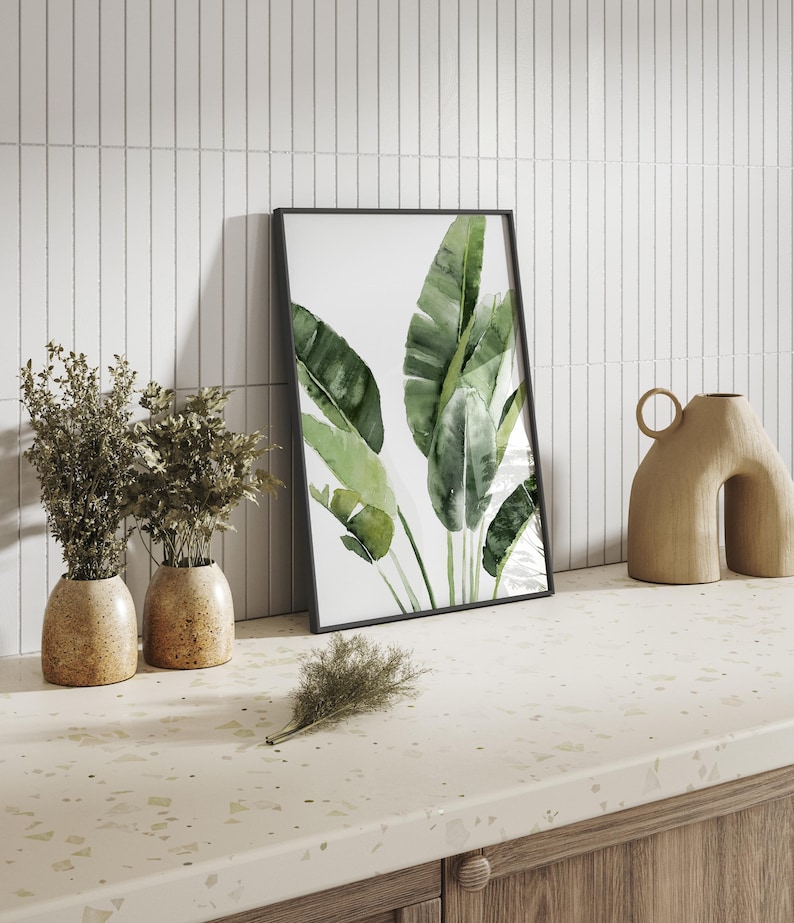 May include: A framed watercolor print of green tropical leaves against a white background. The print is leaning against a white tiled wall, next to two small brown ceramic vases with dried plants. The vases are on a light beige countertop with a speckled pattern.