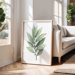 May include: A framed botanical print showing a watercolour illustration of a green leafy branch with small berries. The artwork is displayed in a light wood frame, leaning against a wall near a window and a beige sofa.