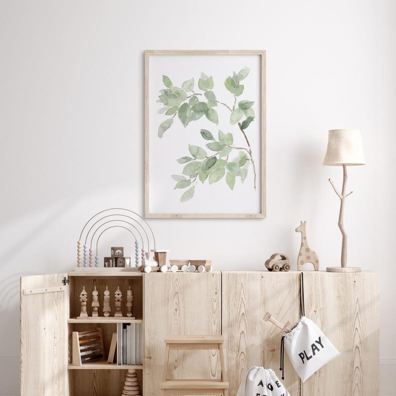May include: A light wood cabinet with two doors and three shelves. The cabinet is decorated with a watercolor print of green leaves in a wooden frame. The cabinet is also decorated with a wooden giraffe figurine, a wooden toy car, and two canvas bags with the words "PLAY" and "A to Z" printed on them.