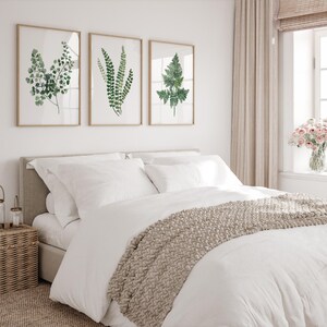 May include: A white bed with a beige knitted throw blanket and three framed botanical prints of ferns on a white wall. The prints are in a simple, modern style.