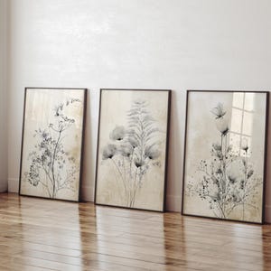 Könnte beinhalten: Drei gerahmte botanische Kunstdrucke. Jeder Druck zeigt ein anderes schwarz-weißes Blumendesign auf beigem Hintergrund. Die Rahmen sind dunkelbraun und lehnen an einer weißen Wand auf einem Holzboden.