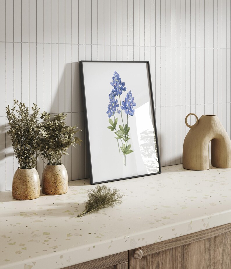 K&ouml;nnte beinhalten: Ein gerahmter Aquarelldruck von Blausternblumen, einer Wildblumenart, die in Texas heimisch ist, vor einem wei&szlig;en Fliesenhintergrund. Zwei Topfpflanzen und eine dekorative Vase stehen auf einer gesprenkelten Arbeitsplatte.