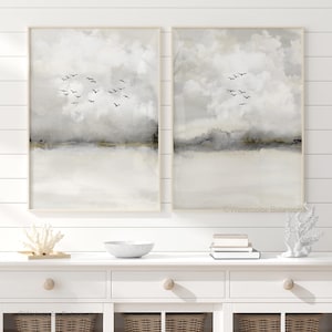 May include: Two framed landscape art prints. Each print features a watercolor design of a cloudy sky with a flock of birds in flight. The artwork has a neutral color palette of white, gray, and gold.