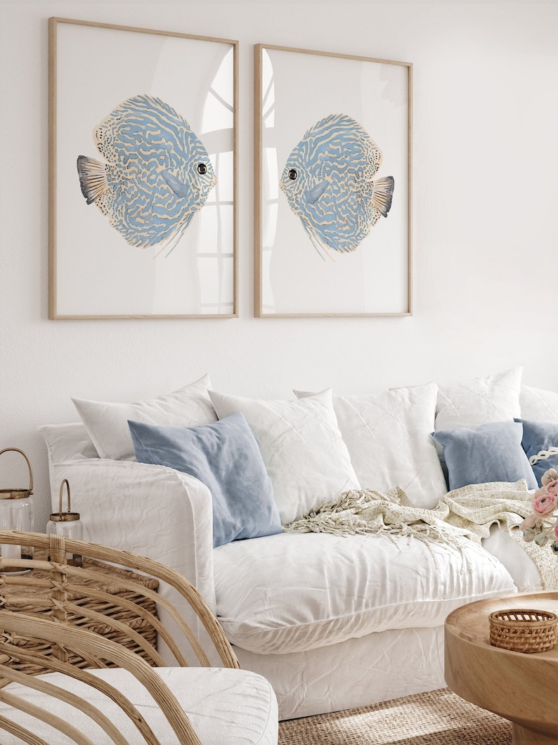 Minimalist coastal wall decor featuring watercolor blue discus fish inserted into wooden frames hangs above a white sofa in a modern coastal living room.