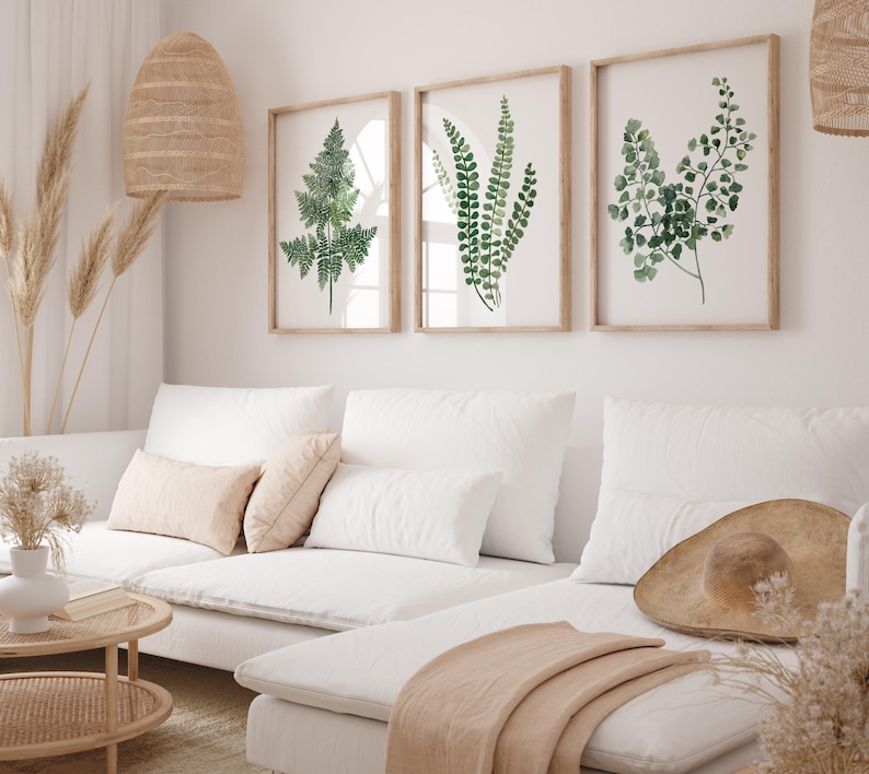 May include: Three framed watercolor prints of ferns in shades of green, hanging on a white wall above a white sectional sofa with a throw blanket and a straw hat.