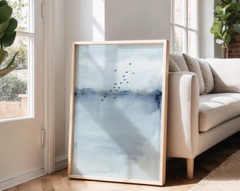 May include: A framed abstract art print featuring a watercolor landscape in shades of blue and gray. A flock of birds flies across the sky. The artwork is displayed in a light wood frame, leaning against a wall near a white sofa.