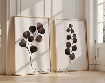 May include: Two framed vertical wall art prints featuring dark brown botanical illustrations of eucalyptus branches on a light cream background. Each print is encased in a light-colored wooden frame with the text Eveline Patrzalek Art.