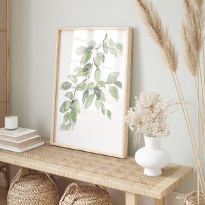 May include: A framed watercolor painting of a green leafy branch on a white background. The painting is hanging on a wall above a wooden bench with a woven rattan top. There are two woven baskets and a white vase with dried flowers on the bench.