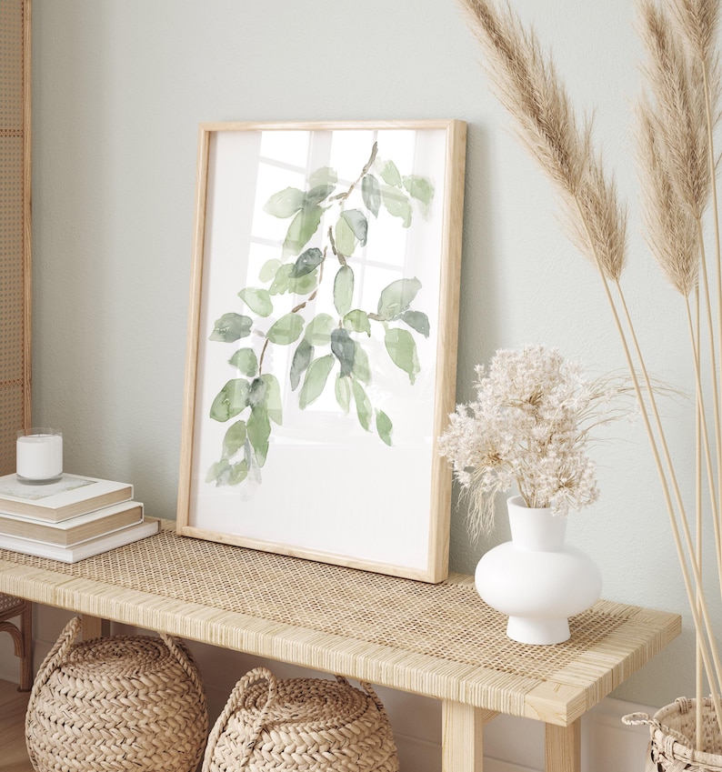 May include: A framed watercolor painting of a green leafy branch on a white background. The painting is hanging on a wall above a wooden bench with a woven rattan top. There are two woven baskets and a white vase with dried flowers on the bench.