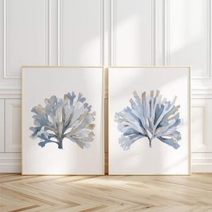 May include: Two vertical rectangular wall art prints featuring watercolor illustrations of sea coral. Each print displays a central branching coral structure in shades of muted blue, grey, and beige against a plain white background.