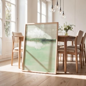 Puede incluir: Una impresión artística enmarcada que presenta un paisaje sereno con una paleta de colores verde y blanco. La obra de arte representa una masa de agua, árboles y una bandada de pájaros contra un cielo nublado. El marco es de madera clara.