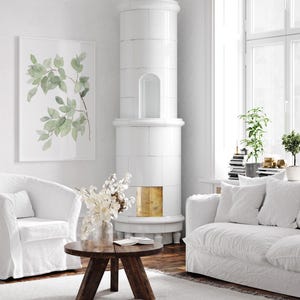 May include: A white living room with a white tiled wood-burning stove, a white couch with white throw pillows, a white armchair, a round wooden coffee table, and a framed watercolor print of green leaves.