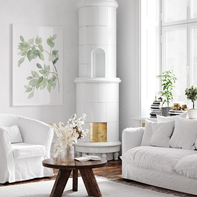 May include: A white living room with a white tiled wood-burning stove, a white couch with white throw pillows, a white armchair, a round wooden coffee table, and a framed watercolor print of green leaves.