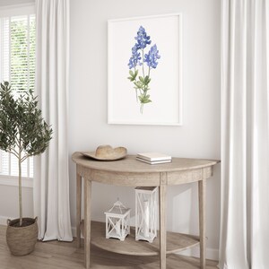 K&ouml;nnte beinhalten: Ein wei&szlig; gerahmter Druck eines Aquarells von blauen Lupinen auf wei&szlig;em Hintergrund. Der Druck h&auml;ngt an einer wei&szlig;en Wand &uuml;ber einem halbmondf&ouml;rmigen Holztisch mit einem Hut, einem Buch und zwei wei&szlig;en Laternen auf dem Tisch.