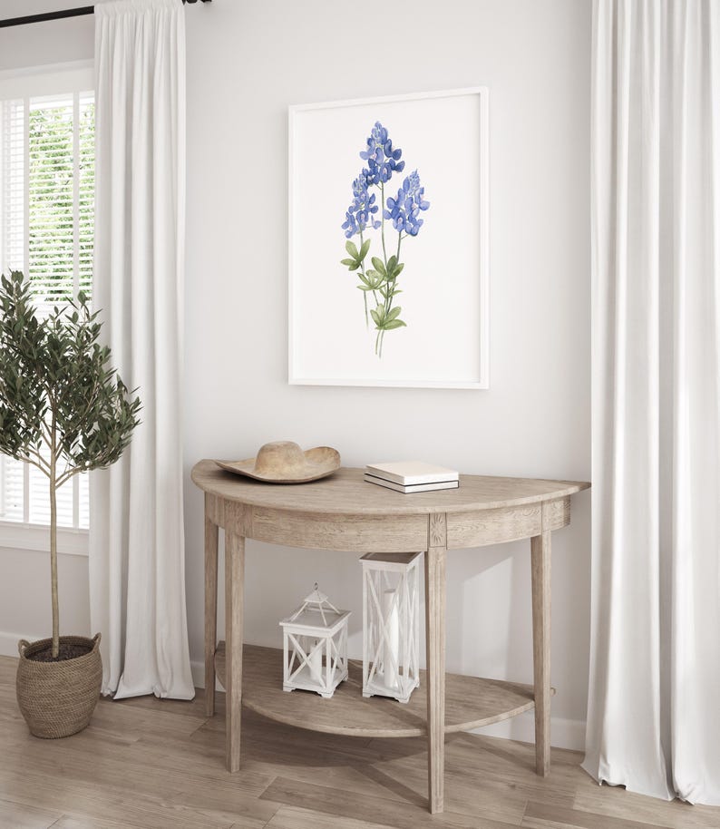 K&ouml;nnte beinhalten: Ein wei&szlig; gerahmter Druck eines Aquarells von blauen Lupinen auf wei&szlig;em Hintergrund. Der Druck h&auml;ngt an einer wei&szlig;en Wand &uuml;ber einem halbmondf&ouml;rmigen Holztisch mit einem Hut, einem Buch und zwei wei&szlig;en Laternen auf dem Tisch.