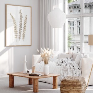 Pu&ograve; includere: Un soggiorno luminoso con una palette di colori neutri. Un tavolino in legno poggia su un tappeto grigio, con un divano bianco e una stampa botanica incorniciata sulla parete. Una lampada a sospensione bianca pende dal soffitto e un pouf intrecciato &egrave; posto sul pavimento. La stanza ha un design minimalista.