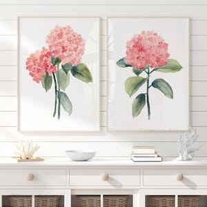 May include: Two framed watercolour paintings of pink hydrangeas with green leaves on a white wood background. The paintings are hung side by side on a white wall.