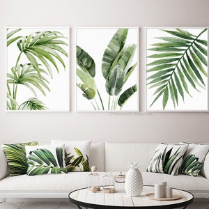 Above a couch hang 3 fine art prints depicting green tropical leaves on a white background. Their size can be customized in centimeters or inches.