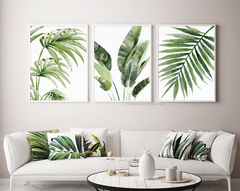 Above a couch hang 3 fine art prints depicting green tropical leaves on a white background. Their size can be customized in centimeters or inches.