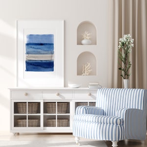 Könnte beinhalten: Ein weißer Schrank mit Glastüren und Weidenkörben. Ein blaugestreifter Sessel steht vor dem Schrank. Ein weißer Rahmen mit einem blauen abstrakten Aquarellbild hängt an der Wand über dem Schrank.