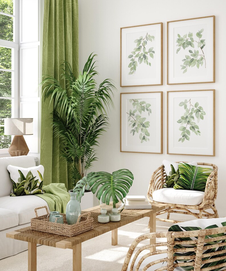 May include: A living room interior design with a white sofa, two wicker chairs with green leaf patterned cushions, a wooden coffee table with a woven tray, a large green potted plant, and four framed watercolor paintings of green leaves on the wall.