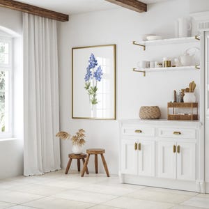 K&ouml;nnte beinhalten: Eine wei&szlig;e K&uuml;che mit einem wei&szlig;en Schrank mit goldenen Beschl&auml;gen, einer wei&szlig;en Arbeitsplatte und zwei Regalen mit goldenen Halterungen. Die Regale sind mit einer Vase, einer Kanne und anderen K&uuml;chenutensilien dekoriert. &Uuml;ber dem Schrank h&auml;ngt ein gerahmtes Bild mit blauen Blumen.