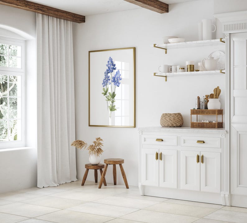 K&ouml;nnte beinhalten: Eine wei&szlig;e K&uuml;che mit einem wei&szlig;en Schrank mit goldenen Beschl&auml;gen, einer wei&szlig;en Arbeitsplatte und zwei Regalen mit goldenen Halterungen. Die Regale sind mit einer Vase, einer Kanne und anderen K&uuml;chenutensilien dekoriert. &Uuml;ber dem Schrank h&auml;ngt ein gerahmtes Bild mit blauen Blumen.