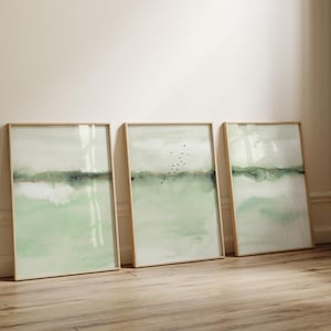 Op de afbeelding: Drie ingelijste abstracte kunstprints. Elke print toont een aquarel landschap in tinten groen, grijs en wit. De middelste print bevat een vlucht zwarte vogels. De lijsten zijn licht van kleur en de kunst leunt tegen een muur.