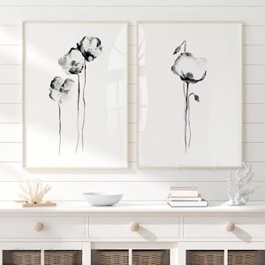 May include: Two framed watercolour prints of black and white poppy flowers. One print features three flowers, the other one flower. The prints are displayed on a white cabinet with decorative items.