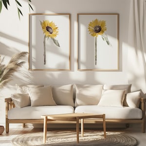 Op de afbeelding: Twee ingelijste aquarelprints van zonnebloemen. Elke print toont een gele zonnebloem met een donkerbruin hart en een groene stengel. De lijsten zijn lichtbruin. De prints worden weergegeven boven een lichtgekleurde bank met beige kussens.