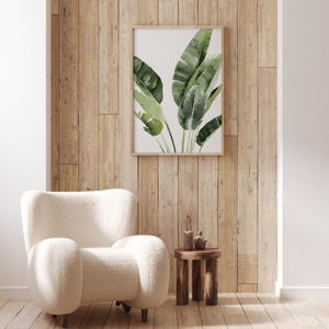May include: A white fluffy armchair with a wooden side table and a framed print of green tropical leaves on a wood panel wall.